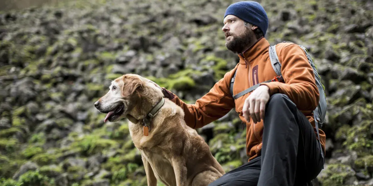 A man in an orange jacket and blue beanie rests during a hiking trip with his golden dog.
