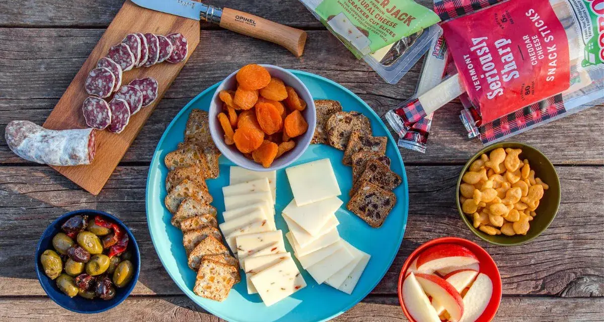 A delicious spread of camping snacks: sliced salami, cheese, crackers, dried apricots, olives, apple slices, and goldfish crackers.