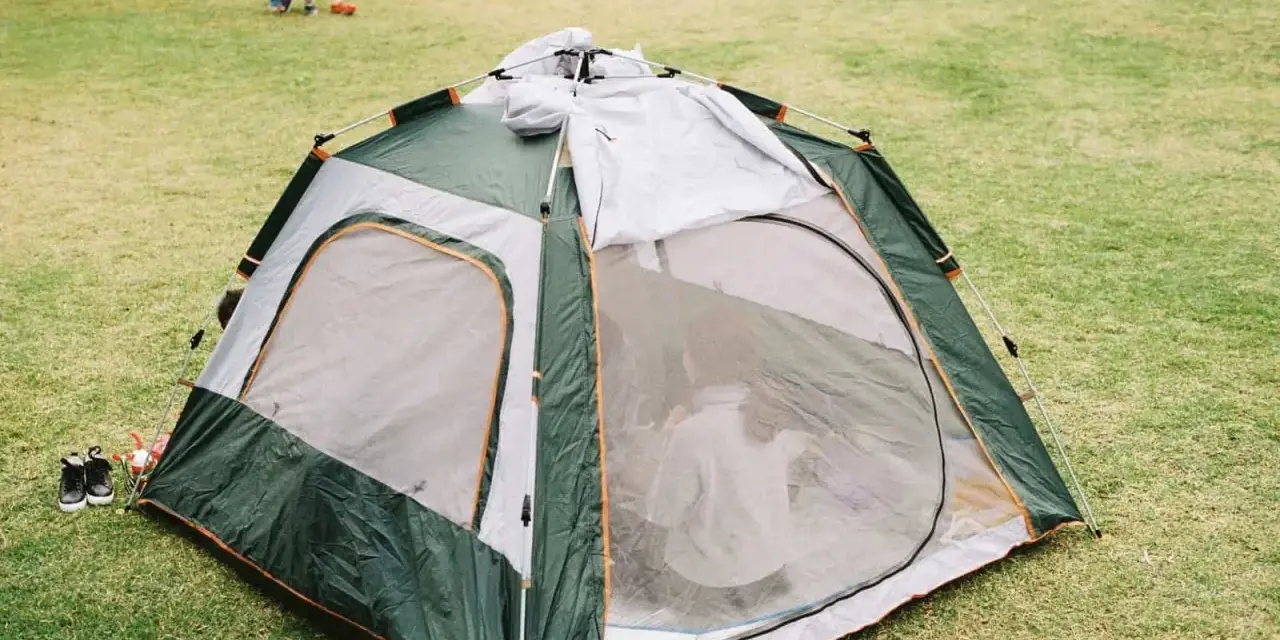 A green and gray tent is partially set up on a grassy field. A child plays in the background, learning how to set up a tent.