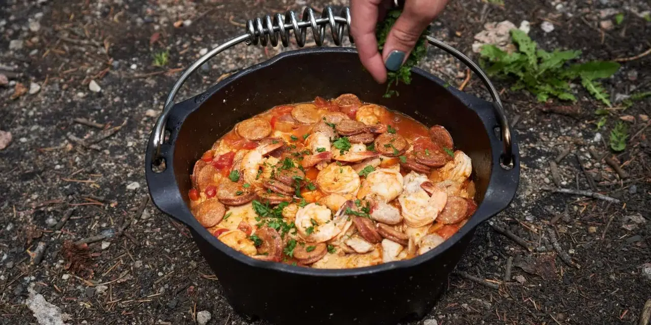 A hand garnishes a hearty dutch oven paella with fresh parsley. This campfire meal features shrimp, sausage, and rice in a rich tomato sauce.