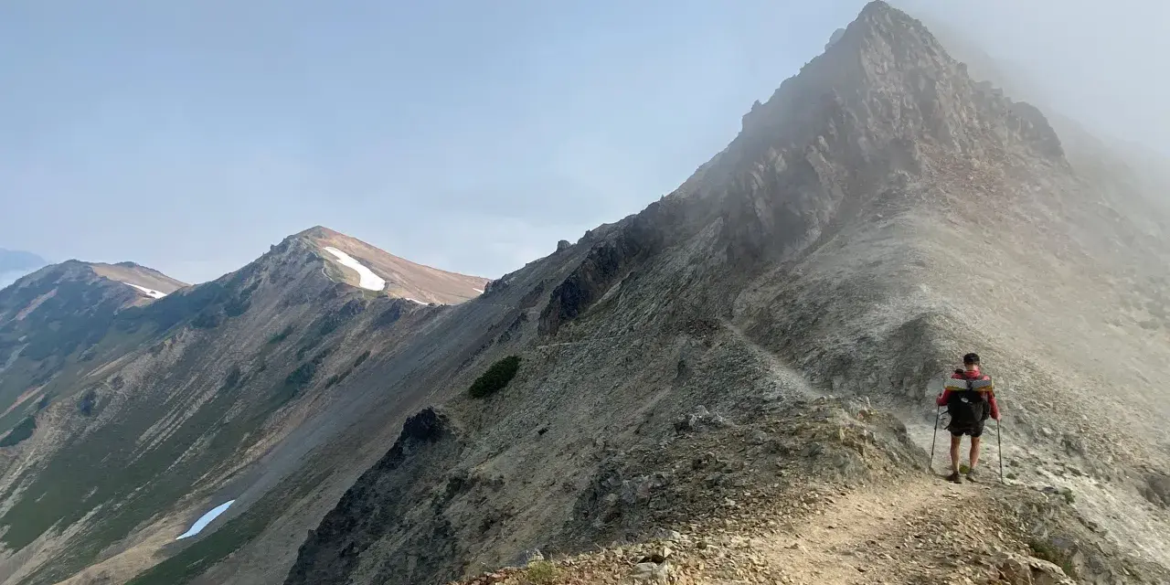 A hiker with a backpack and trekking poles walks along a narrow mountain ridge, a scenic path to learn how to get better at hiking.