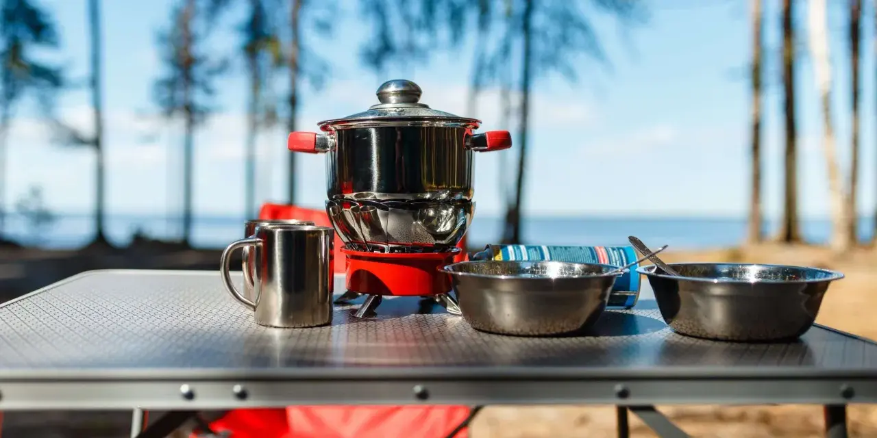 A complete camping kitchen setup with a stove, pots, bowls, and mugs on a table, ready for outdoor cooking by a lake.