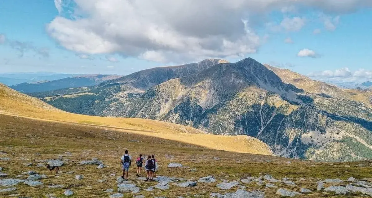 Hikers with backpacks traverse a rocky, grassy meadow on a pyrenees hut to hut hiking adventure, with majestic mountains under a cloudy sky.