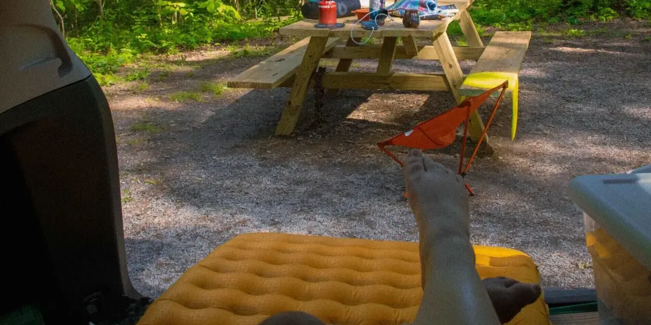A person's legs rest on an orange sleeping pad inside a car. A picnic table with camping gear sits in a wooded area, ready for a road trip checklist.