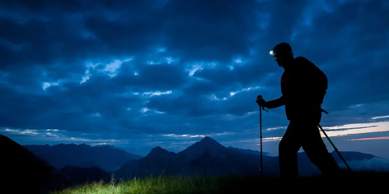 Silhouette of a hiker with a headlamp on, trekking poles in hand, on a night hike through a dramatic, cloudy sky over mountains.