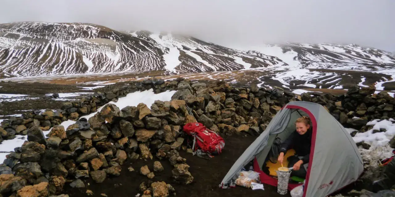 A hiker prepares a meal inside a tent at one of the Laugavegur trail huts, surrounded by snow-dusted mountains and a stone wall.