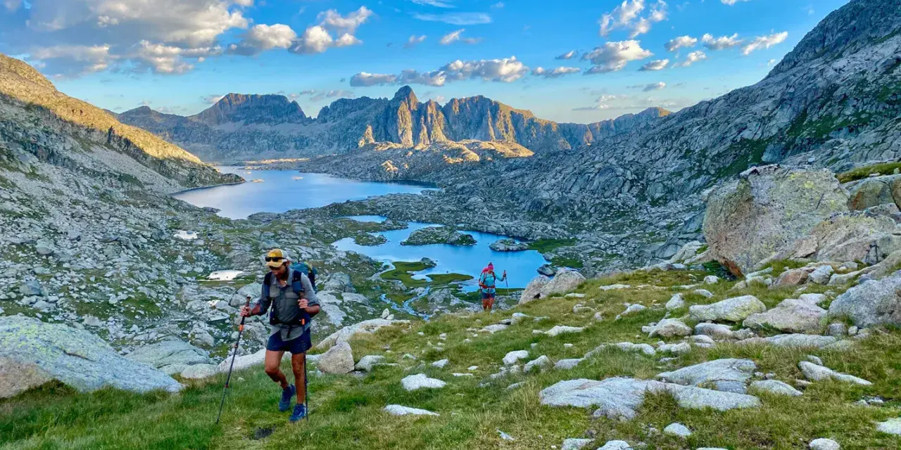 Hikers ascend a grassy slope on the HRP trail, with a stunning mountain lake and jagged peaks in the background under a cloudy sky.