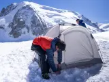 Two people set up a tent for winter camping amidst a snowy mountain landscape under a clear blue sky.