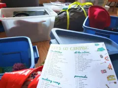 A "Let's Go Camping" checklist is spread out among blue and white storage bins, ready for a trip.