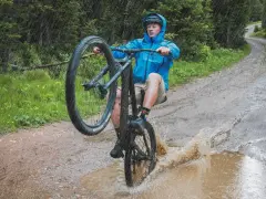 A mountain biker in a blue jacket performs a wheelie through a large puddle, splashing water as it rains heavily on a forest trail.