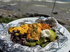 Delicious campfire chicken foil packets with corn, black beans, rice, jalapeños, and a lime wedge, ready to be enjoyed by the water.