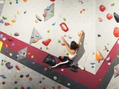 A woman is bouldering rock climbing, reaching for an orange hold on a colorful, textured wall.