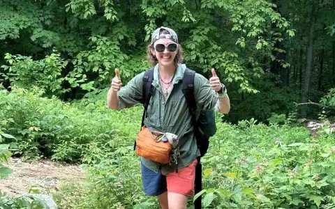 A hiker in a green, breathable hiking shirt material, gives a double thumbs-up in a lush forest.