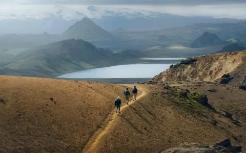 Hikers traverse a golden path on a mountain ridge, with a vast, serene lake and snow-capped peaks in the distance. This iconic landscape hints at the epic scale of how long is the Laugavegur trail.