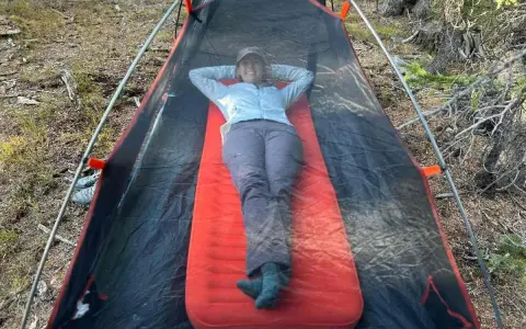 A person relaxes inside a tent on a bright orange Therm-a-Rest NeoAir XLite sleeping pad, enjoying the outdoors.
