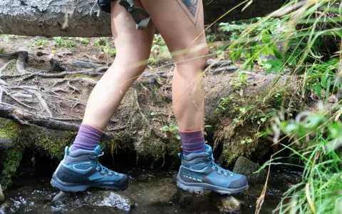 A hiker steps through a stream, testing how to waterproof hiking boots. Their blue boots and purple socks are ready for any wet trail.