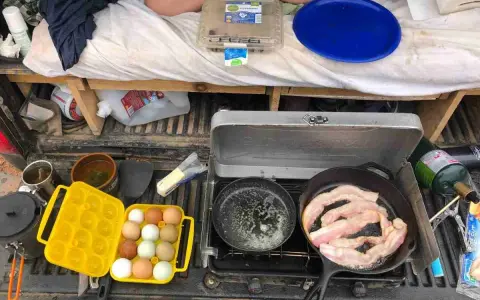 A camp kitchen checklist in action: bacon sizzles in a cast-iron pan, eggs are ready in a yellow carrier, and a portable stove is set up.