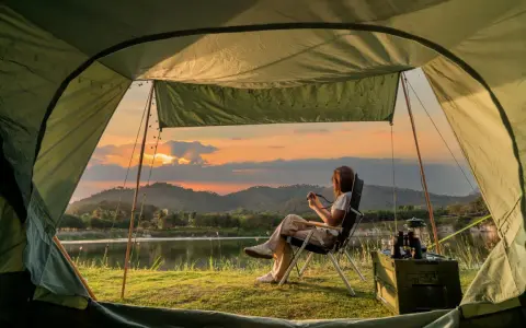 A woman enjoys solo camping by a lake at sunset, holding a camera. The tent offers a cozy view of the serene landscape.