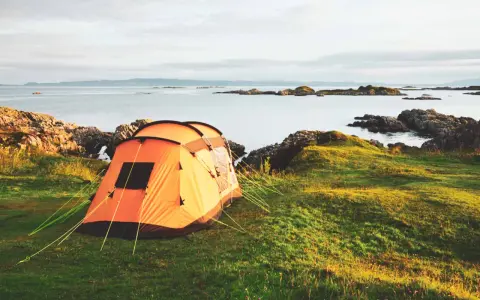 An orange tent is set up on a grassy hill overlooking a calm sea with rocky islands. This is a perfect spot for tent camp sites.