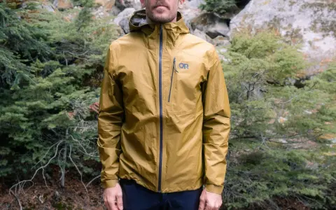 Man wearing a golden ultralight waterproof jacket with an OR logo, standing in a forest.