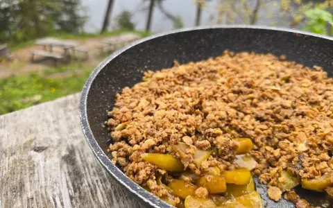 A warm, bubbling campfire apple crisp in a pan, ready to be enjoyed outdoors by a lake.