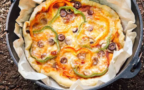 A delicious dutch oven pizza camping treat, topped with melted cheese, colorful bell peppers, and olives, baked to perfection on parchment paper.