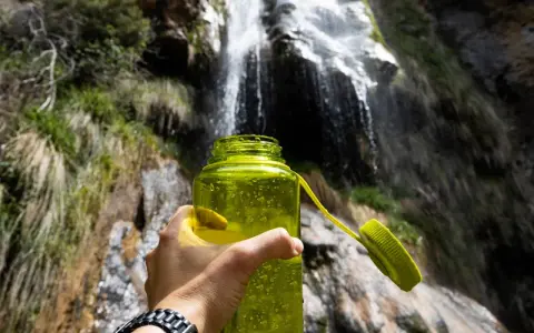 A hand holds a bright green water bottle, ready for a refreshing drink after a long hike. Behind it, a beautiful waterfall cascades down a rocky cliff, surrounded by lush greenery.