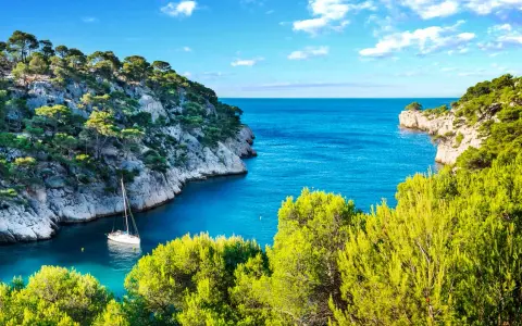 A sailboat anchors in a serene cove, surrounded by lush green trees and dramatic cliffs. This idyllic scene evokes the feeling of a perfect camping trip in Cassis.
