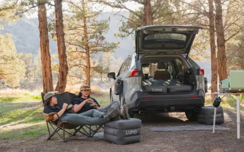 Two people enjoy a meal in camping chairs next to a car with an open trunk, showcasing clever car camping hacks with an inflatable mattress and a portable stove.