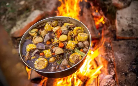 A pot of hearty camping meals, filled with corn, potatoes, carrots, and meat, simmers over a crackling campfire.