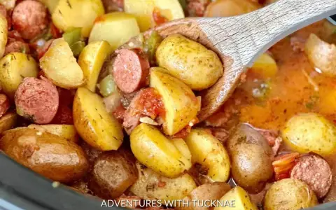 A wooden spoon scoops up hearty potatoes and sausage from a crockpot, perfect for camping crockpot meals.
