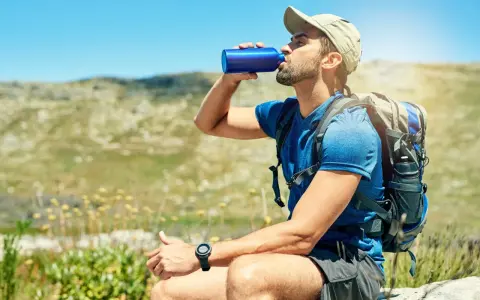 A hiker takes a refreshing drink from a blue water bottle, demonstrating how to stay hydrated while hiking in a sunny, mountainous landscape.