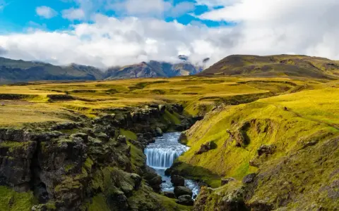 A winding path on the Laugavegur trail in Iceland leads past a mossy canyon with a cascading waterfall.