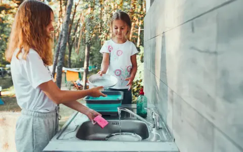 Two girls are washing dishes camping. One holds a pan over a sink while the other prepares to rinse it.