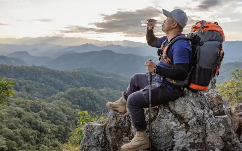 A hiker drinks water on a mountain peak, contemplating how much water to take backpacking.