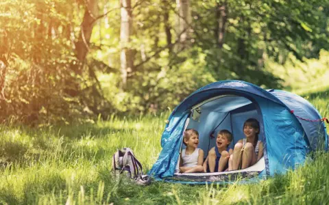 Three children laugh inside a blue tent, enjoying camping activities in a sun-dappled forest.