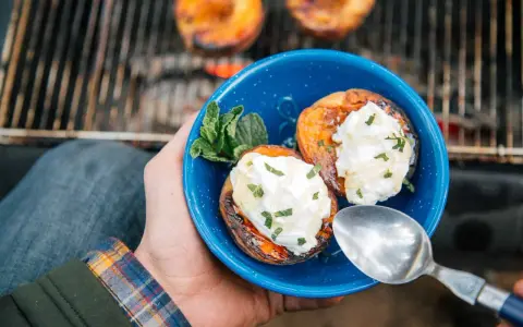 A hand holds a blue bowl with two grilled peaches topped with cream and mint. Campfire peaches are a perfect dessert.