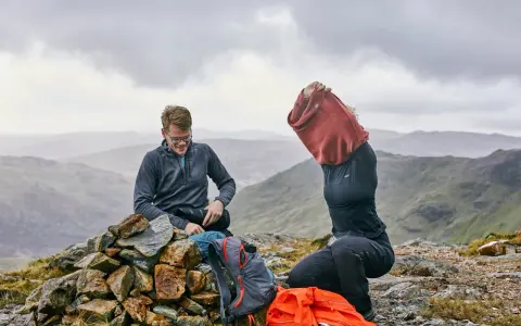 Hikers on a rocky summit adjust their backpacking clothes. One man smiles while sitting on a cairn, next to backpacks. Another person pulls on a red sweater.