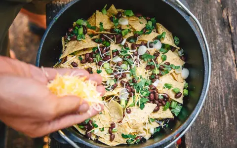 Making delicious camping nachos in a Dutch oven, adding shredded cheese to tortilla chips, black beans, and green onions.