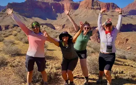 Four women pose with arms raised in a desert canyon, dressed for hiking in hot weather with shorts and light tops.