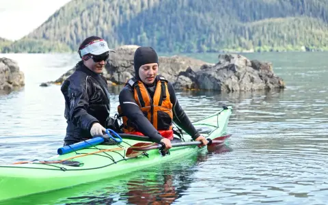 Two kayakers in a bright green kayak practice a paddle float re-entry in calm water, with a forested mountain in the background.