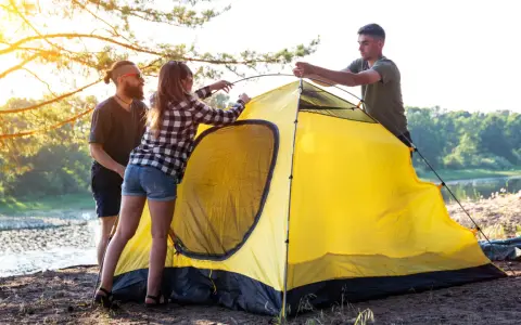 Friends setting up a bright yellow tent by a lake, learning how to choose a campsite with a beautiful sunset.