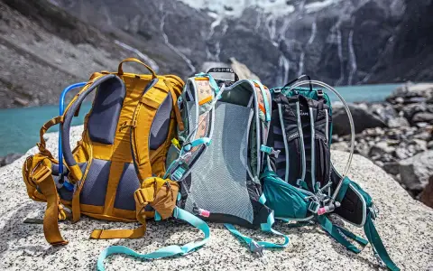 Three Osprey backpacks sit on rocks by a lake, ready for a hike. Learn how to pack a day pack for hiking with these essential tips.
