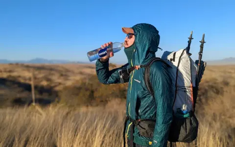 A hiker takes a refreshing drink from a Smartwater bottle while carrying a large backpack and trekking poles.