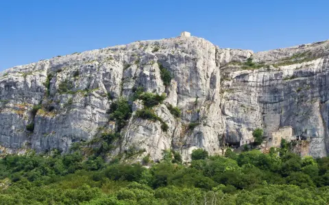 A majestic cliff face rises above a lush green forest, with a small building nestled into the rock at Sainte Baume.