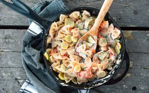 A hearty camping pasta dish with farfalle, zucchini, and tomatoes in a creamy sauce, served in a cast-iron skillet.
