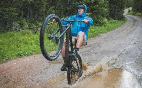 A mountain biker in a blue jacket performs a wheelie through a large puddle, splashing water as it rains heavily on a forest trail.