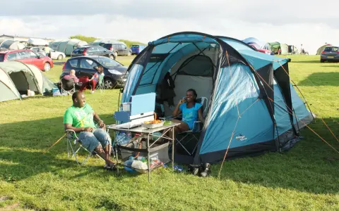 A couple enjoys a meal at their campsite, a perfect example of how to camp. They sit in chairs beside a blue tent, with food laid out on a portable table.