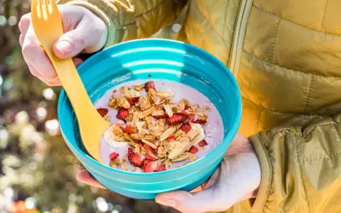 A person in a yellow jacket holds a blue bowl filled with a pink smoothie topped with granola, dried strawberries, and banana slices. Perfect for camping breakfast ideas.