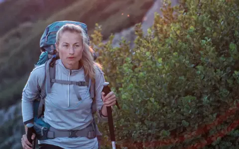 A woman with blonde hair, wearing a grey jacket and a large backpack, is backpacking hiking up a rocky, vegetated slope. She holds a trekking pole.